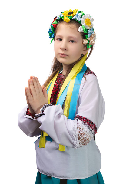 Ένα κοριτσάκι με εθνικά ουκρανικά ρούχα με δακρυσμένα μάτια δίπλωσε τα χέρια της μπροστά της και προσεύχεται για το τέλος του πολέμου στην Ουκρανία. Φωτογραφία στούντιο σε λευκό φόντο. - Φωτογραφία, εικόνα