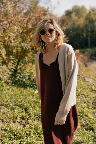 Прекрасная очаровательная женщина с волнистыми светлыми волосами в виноградном платье и бежевом свитере в солнечных очках позирует перед камерой на фоне осеннего леса. Высокое качество фото - Фото, изображение
