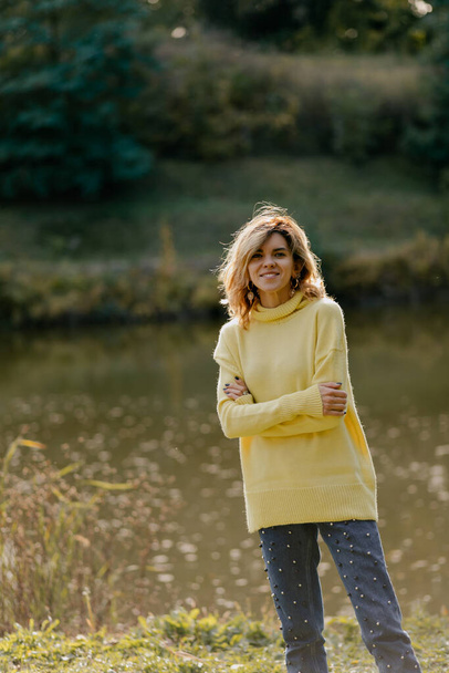 encantadora dama sonriente en suéter amarillo con pelo claro sonriendo a la cámara mientras camina por el lago con vista al bosque en un día soleado. Foto de alta calidad - Foto, Imagen