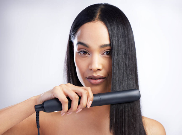 How do I get straight hair, you ask. Shot of an attractive young woman standing alone in the studio and styling her hair with a hair straightener. - Photo, image