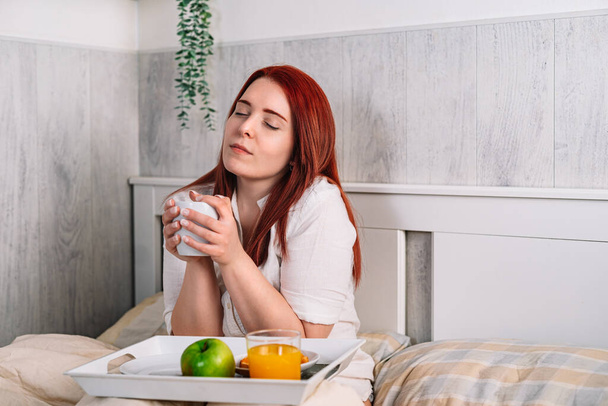 сексуальна руда молода жінка сидить у ліжку сніданок, вона відпочиває з чашкою кави в руках
. - Фото, зображення