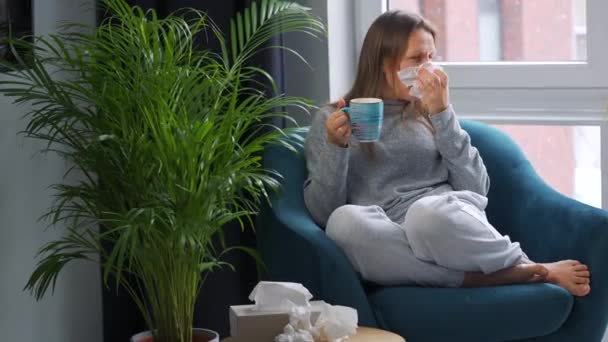 Epäterve nainen istuu tuolissa, juo kuumaa teetä tai flunssalääkettä ja aivastaa tai puhaltaa nenänsä lautasliinaan, koska hänellä on kylmä, flunssa, sepelvaltimovirus. Ulkona sataa lunta. - Materiaali, video