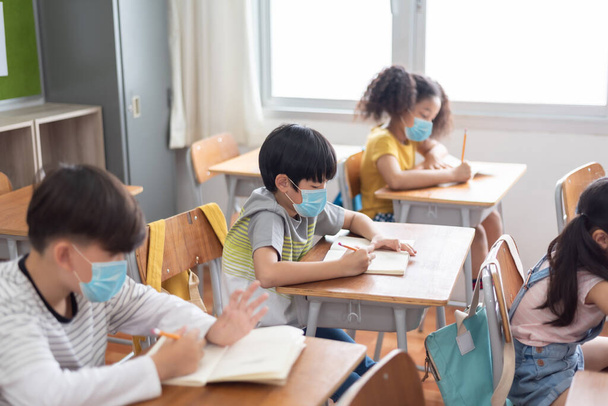 Skoncentrowany Zróżnicowany pochodzenie etniczne małych dzieci szkolnych noszących ochronną maskę na twarzy siedzących przy biurku w klasie, piszących. - Zdjęcie, obraz