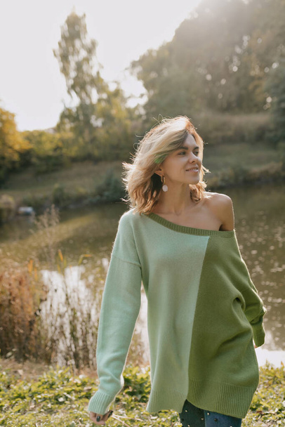 Spettacolare ragazza elegante con i capelli chiari indossando maglione verde con spalla nuda sta camminando nel parco verde con sfondo lago al sole. Foto di alta qualità - Foto, immagini