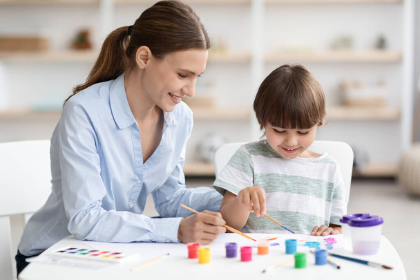 Χαριτωμένο αγοράκι και η νεαρή μητέρα του ζωγραφίζουν μαζί με πολύχρωμες μπογιές, κάθονται στην τάξη και χαμογελούν - Φωτογραφία, εικόνα