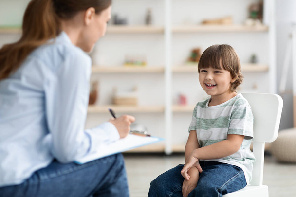 Nahaufnahme Porträt des netten, fröhlichen kleinen Jungen, der mit einer professionellen Kindertherapeutin spricht und ihr eine lustige Geschichte erzählt - Foto, Bild