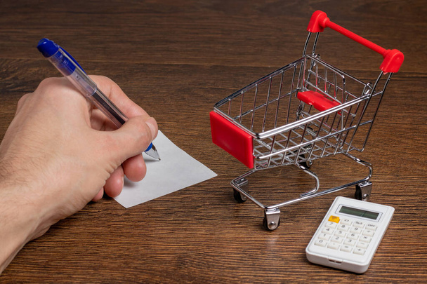 Man calculates the budget. Shopping cart a on table with calculator and paper. Budget of poor and low income family. Rising food and grocery store prices and expensive daily consumer goods concept - Photo, Image