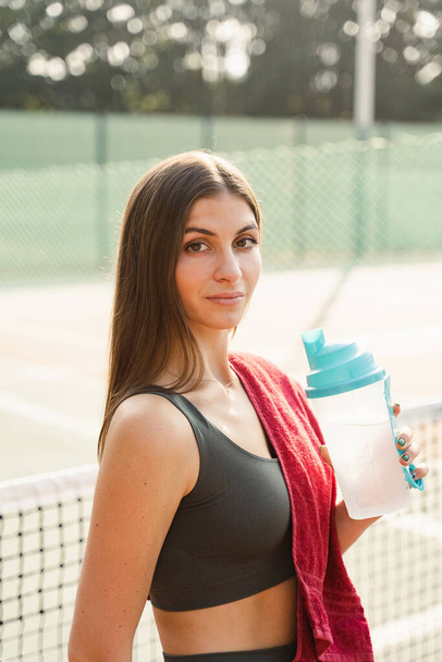 Fit sports model is resting after training and drinking water from a bottle. Sports lifestyle. Beautiful girl posing on the tennis court - Фото, изображение