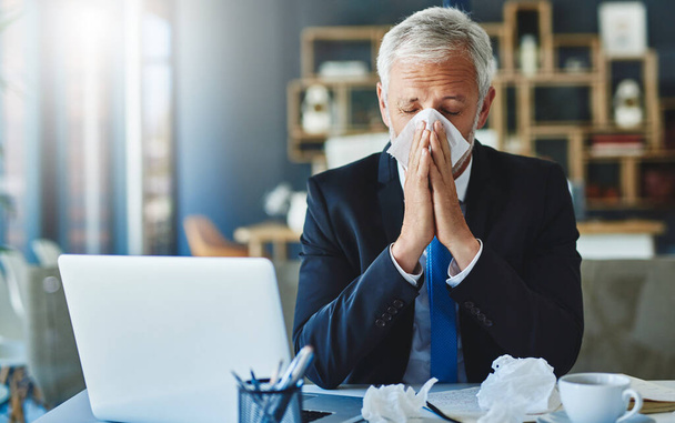 Got to stock up on tissues. Shot of a frustrated businessman using a tissue to sneeze in while being seated in the office. - Photo, Image