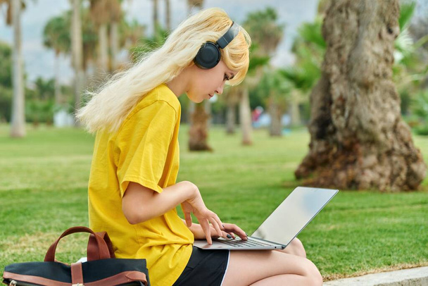 Девушка-подросток в наушниках сидит в парке с ноутбуком. - Фото, изображение