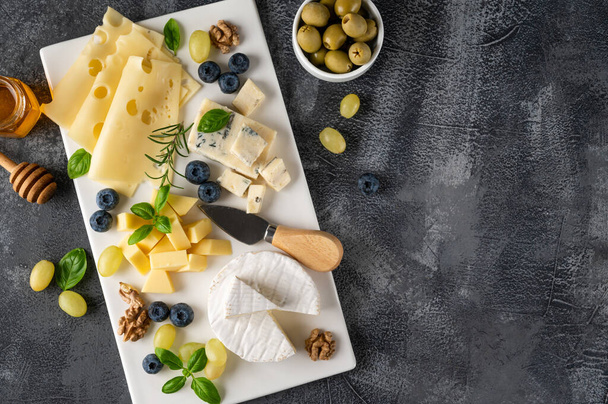 Prato de queijo com uma variedade de queijos, mel, uvas, nozes, azeitonas, mirtilos e ervas frescas em um fundo de concreto. Um lanche festivo - Foto, Imagem