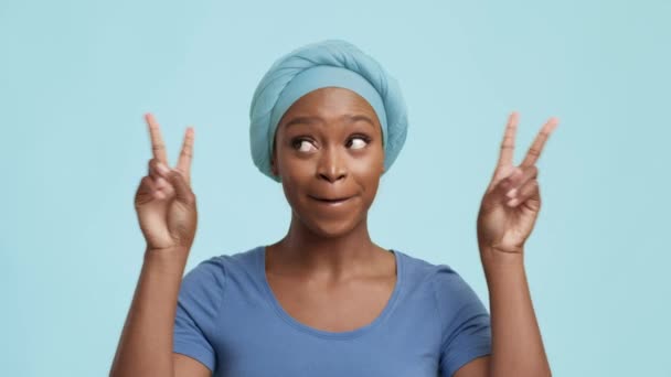 Fröhliche Afroamerikanerin gestikuliert mit V-Zeichen über blauem Hintergrund - Filmmaterial, Video