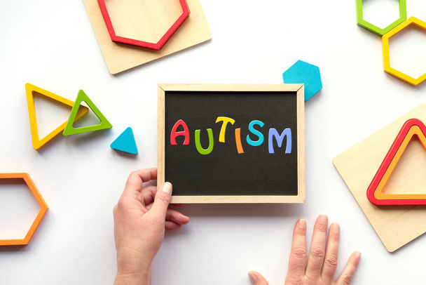 Text Autism, word on blackboard in rainbow colors. Nested wood triangles and hexagons in various bold colors. Chalk board in human hand. Concept flat lay on white. - Fotografie, Obrázek