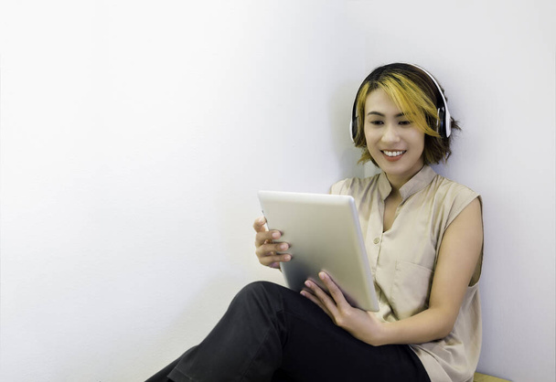 Happy Asian kobieta krótkie włosy w beżowej koszuli bez rękawów patrząc na tabletkę i przy użyciu go z białym bezprzewodowym słuchawką na białym tle ściany z miejsca do kopiowania. Oglądanie filmów, słuchanie koncepcji muzyki. - Zdjęcie, obraz