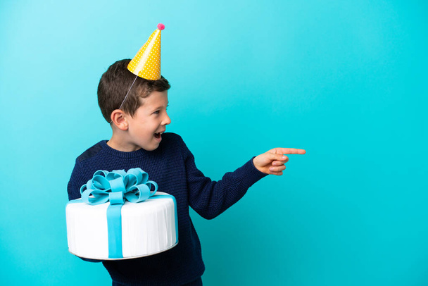 Маленький хлопчик тримає торт на день народження ізольований на синьому фоні, вказуючи пальцем на бік і представляючи продукт
 - Фото, зображення
