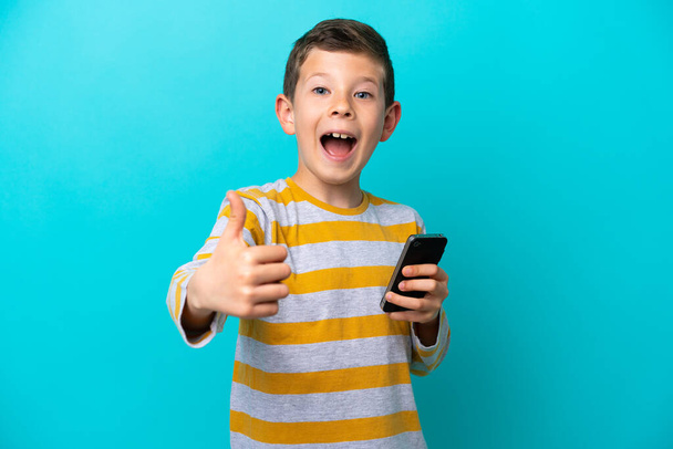 Little boy isolated on blue background using mobile phone while doing thumbs up - Photo, image