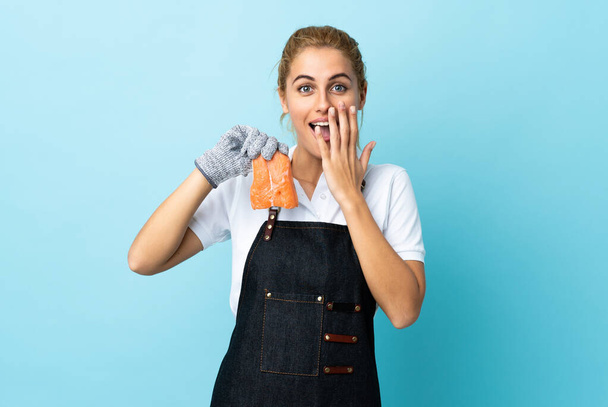 Fishmonger wearing an apron and holding a raw fish over isolated blue background with surprise facial expression - Фото, зображення
