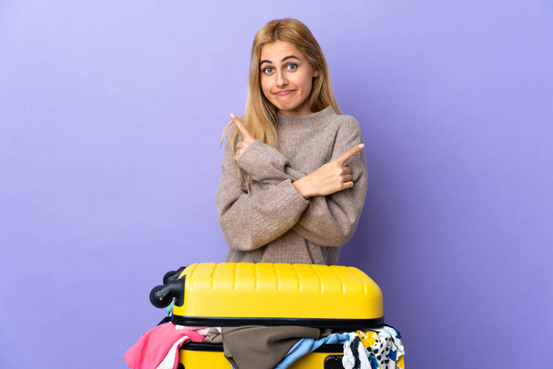 Nuori Uruguayn blondi nainen matkalaukku täynnä vaatteita yli eristetty violetti seinä osoittaa laterals ottaa epäilyksiä - Valokuva, kuva