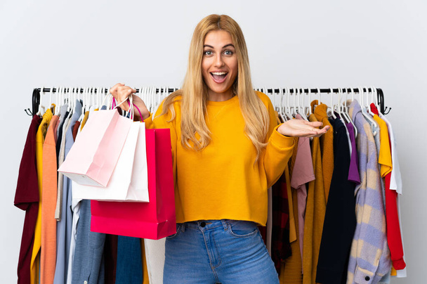 Giovane donna bionda uruguaiana in un negozio di abbigliamento e tenendo borse della spesa con espressione facciale scioccata - Foto, immagini