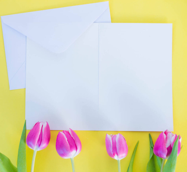white card and envelope on yellow ground with pink tulips with space for text - Valokuva, kuva