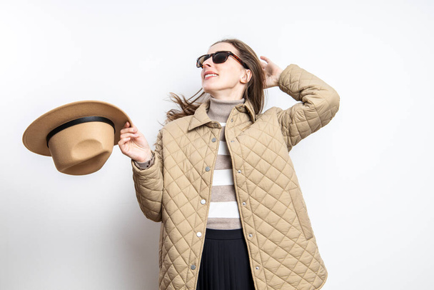 Una joven alegre con gafas y una chaqueta se quitó el sombrero cerca de una pared blanca - Foto, Imagen