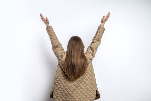 Giovane donna con i capelli lunghi in una giacca alzando le mani vicino a un muro bianco - Foto, immagini