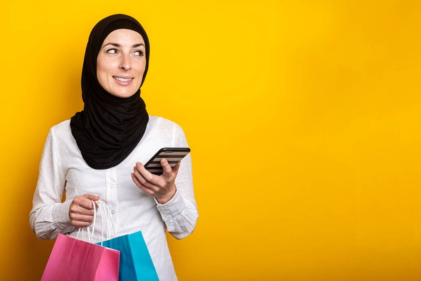 La joven musulmana en hiyab sostiene un teléfono y bolsas de compras sobre un fondo amarillo. Banner. - Foto, imagen