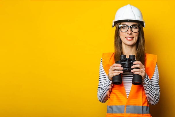 Mujer joven con una sonrisa en un chaleco y un casco sostiene los prismáticos sobre un fondo amarillo. Concepto de construcción, edificio nuevo. Banner. - Foto, Imagen