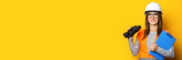 Молодая женщина с улыбкой в жилете и шлеме держит планшет и бинокль на желтом фоне. Баннер. - Фото, изображение