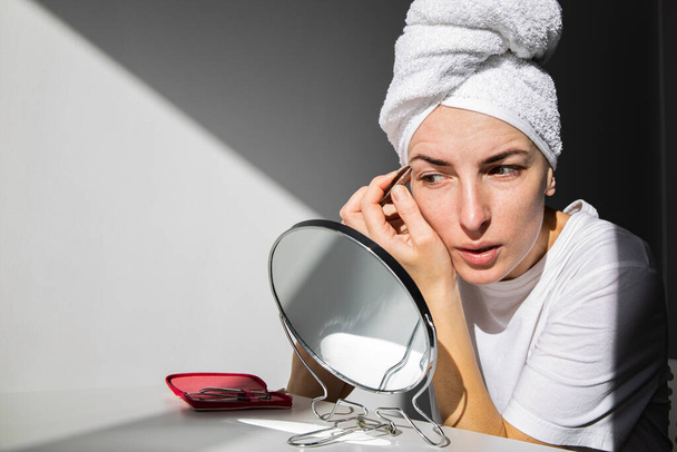Junge Frau ohne Make-up mit Handtuch auf dem Kopf zupft sich die Augenbrauen. - Foto, Bild