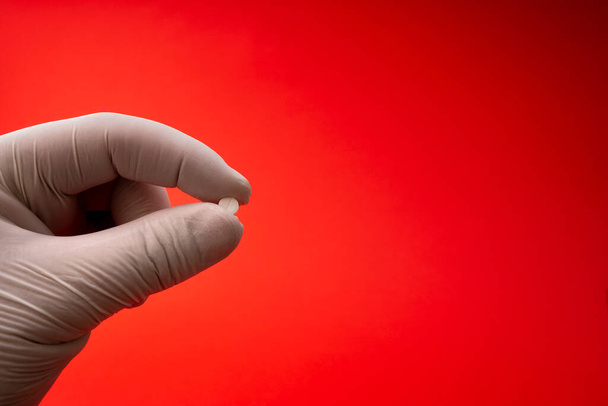 Χέρι στο γάντι λατέξ κρατώντας χάπι απομονώνονται σε κόκκινο φόντο - Φωτογραφία, εικόνα