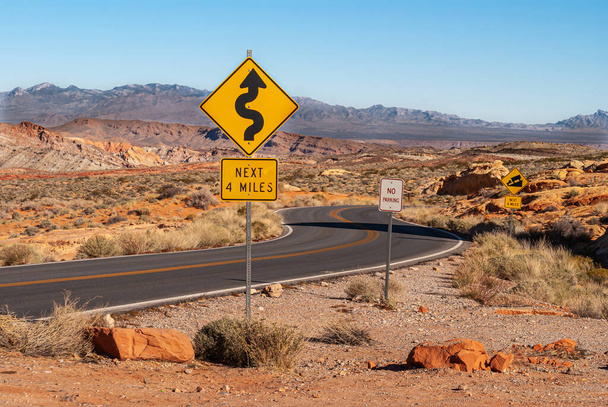 Overton, Nevada, USA - 11 grudnia 2010: Dolina Ognia. Czarny asfalt droga meanders przez szeroki brązowy górski krajobraz pustyni z żółtymi ostrymi zakrętami znak drogowy z przodu. - Zdjęcie, obraz