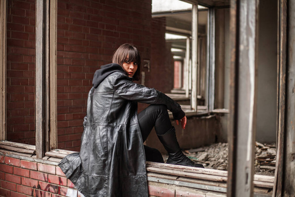 Young woman in a leather coat alone in a destroyed residential building, loneliness and despair in an abandoned city - Valokuva, kuva
