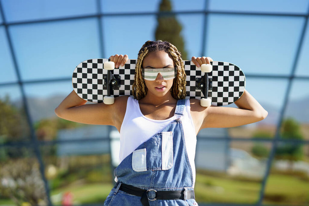 Zwarte vrouw gekleed casual, kijken naar camera met moderne zonnebril en een skateboard. - Foto, afbeelding
