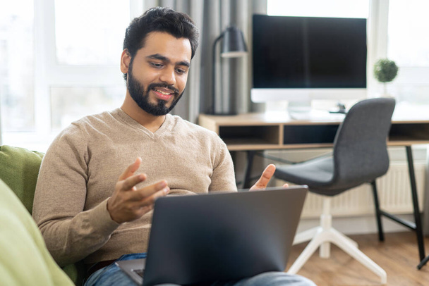 Cheerful middle eastern man in casual wear is sitting on the comfortable sofa and using trendy laptop for video call, talking emotionally with online interlocutor - 写真・画像