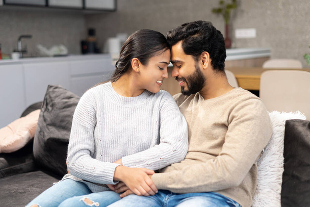 Happy Indian newlyweds moved in new apartment, sitting at the sofa in embraces, looking at each other. Multi ethnic couple in love rent a house together - Fotografie, Obrázek