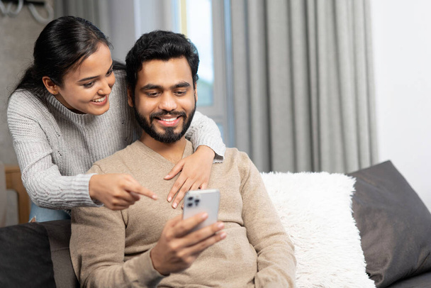 Cheerful couple spends time with smartphone at home. Wife and a husband looks on the screen with smiles sitting on the couch at cozy living room, watching videos - Fotografie, Obrázek