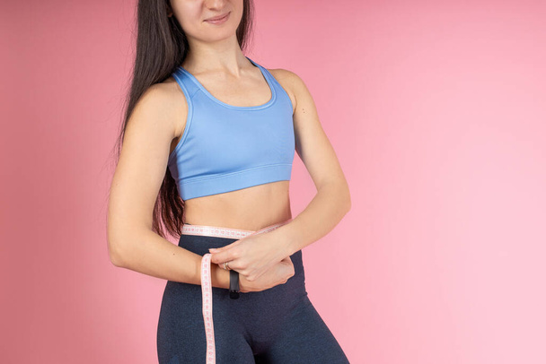 Kouluttajalla on kädessään mittanauha ruumiin mittaamiseksi. Houkutteleva brunette nainen muodikas urheiluvaatteet vaaleanpunainen tausta. Terveiden elämäntapojen käsite - Valokuva, kuva