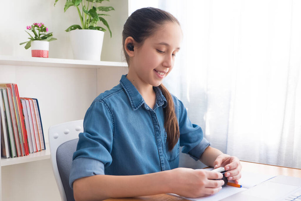 Brunette tiener in een denim shirt maakt gebruik van draadloze koptelefoon om thuis te studeren, studeert, doet huiswerk communiceert op het internet. - Foto, afbeelding