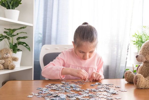 6-7 yaşlarındaki pembe tişörtlü küçük kız evde oyuncak ayıyla bir masada oturuyor ve bulmacaları topluyor.. - Fotoğraf, Görsel