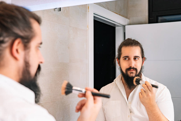 Jongeman die make-up voor de spiegel aanbrengt. Dagelijkse make-up routine in de badkamer - Foto, afbeelding