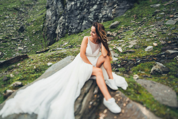Bride with long hair sitting on a stone against the background of rocks and cliffs, and looking to one side. Fashion model. - Foto, afbeelding