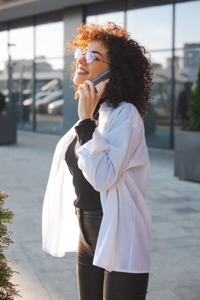 Kiharatukkainen valkoihoinen nainen puhuu matkapuhelimella ulkona käyttäen nykyaikaisia vempaimia viestintään ja työhön. Hymyilevät kasvot. Sivunäkymä Elämäntapa suunnittelu - Valokuva, kuva