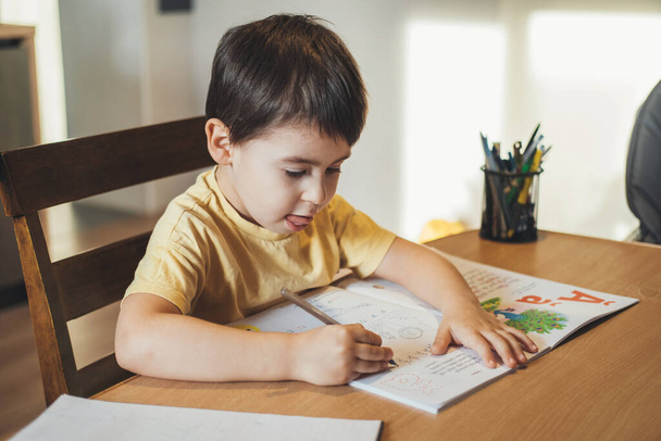 Αγόρι κάνει έκφραση προσώπου μελετώντας σκληρά καθώς κάνει τα μαθήματά του. Πορτρέτο του αγοριού που γράφει στο τραπέζι. Εκπαίδευση των παιδιών. - Φωτογραφία, εικόνα