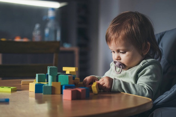 Pequena menina sentada na cadeira alta enquanto brincava com blocos de brinquedo de madeira em casa. Retrato para design de estilo de vida. Desenvolvimento do bebê. - Foto, Imagem
