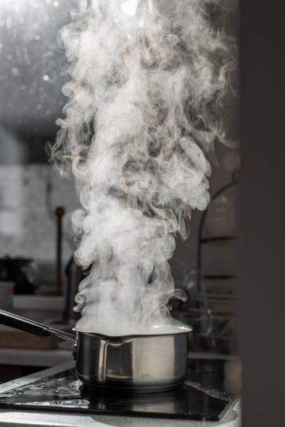 Boiling water with steam in a pot on an electric stove in the kitchen. Blurred background, selective focus. - Photo, image