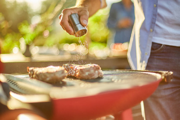 Close up on man's hand seasoning meat on the gas grill on barbecue grill outdoor in the backyard, grilled roasted steak meat, summer family picnic, food on the nature. - Photo, Image