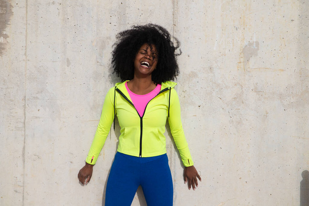 Beautiful young Afro-American woman in bright green and pink sportswear on a grey concrete wall texture background. Woman makes different expressions. Laughing, serious, happy, sad, thinking - Photo, Image