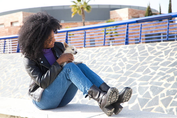 Νεαρή και όμορφη Αφρο-Αμερικανίδα που κάθεται στο πάρκο ενώ παίζει με τον μικρό της σκύλο. Η γυναίκα φοράει καθημερινά και άνετα ρούχα. Κατοικίδια και κατοικίδια ζώα έννοια - Φωτογραφία, εικόνα