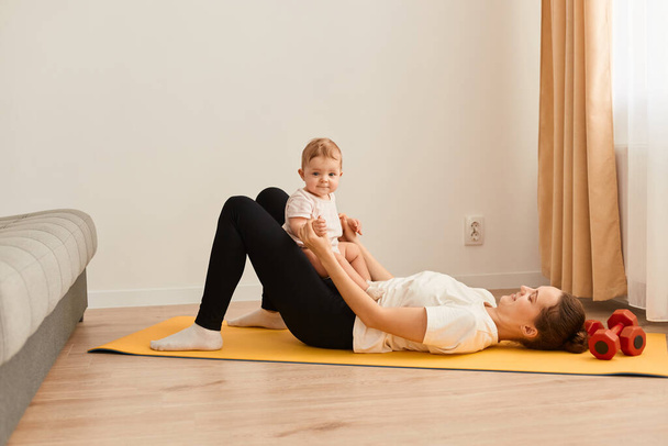 Image of adorable woman wearing white t shirt and black leggins doing sport exercises with baby daughter, having fun with her kid while having workout. - Photo, Image